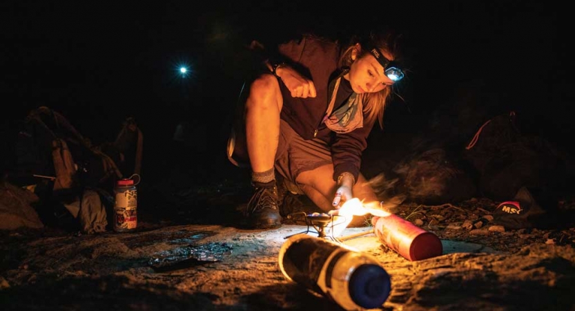 A person wearing a headlamp blows on a small campfire. 